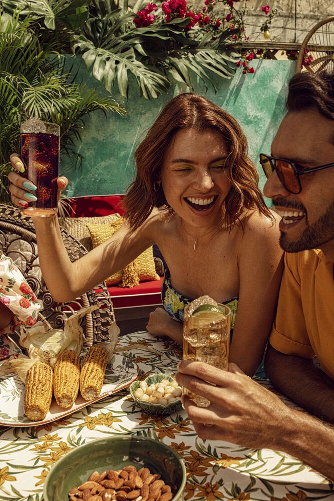 Two people enjoying cocktails and snacks at an outdoor table with green plants.