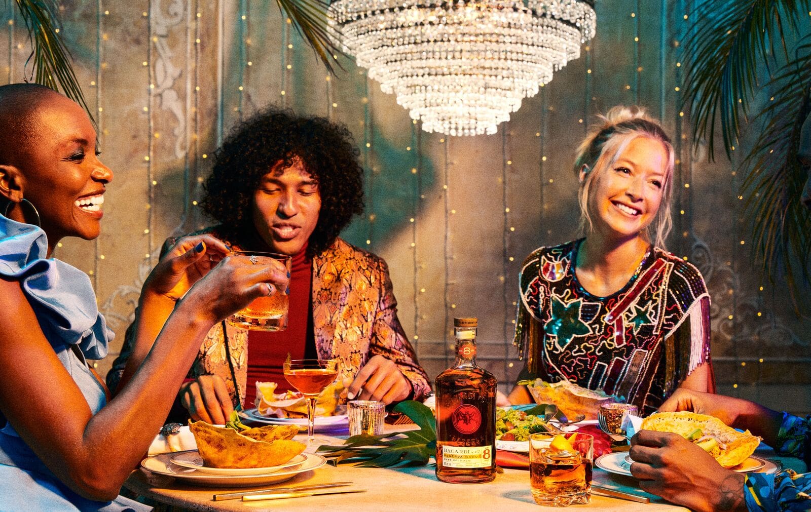 Three people seated at a table with BACARDÍ rum bottle, cocktails, and food, creating a social gathering scene.