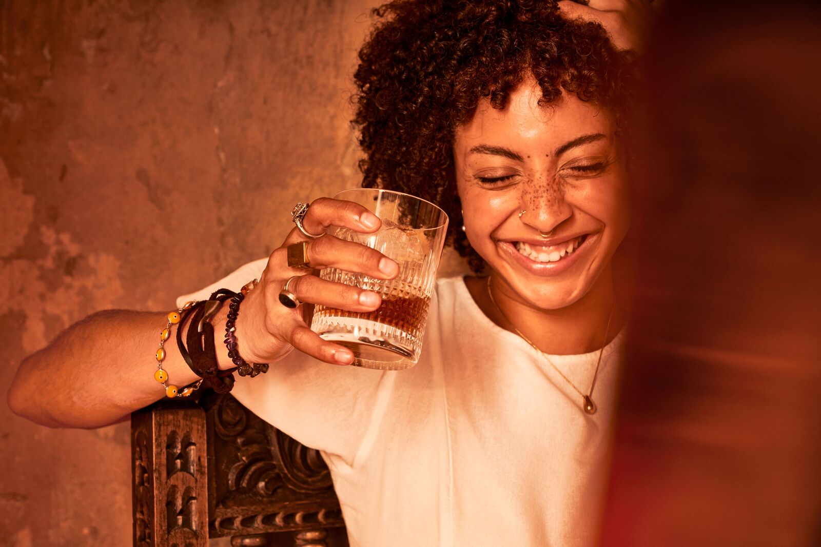 Person smiling with eyes closed, holding a glass of amber liquid while leaning on a wooden chair.