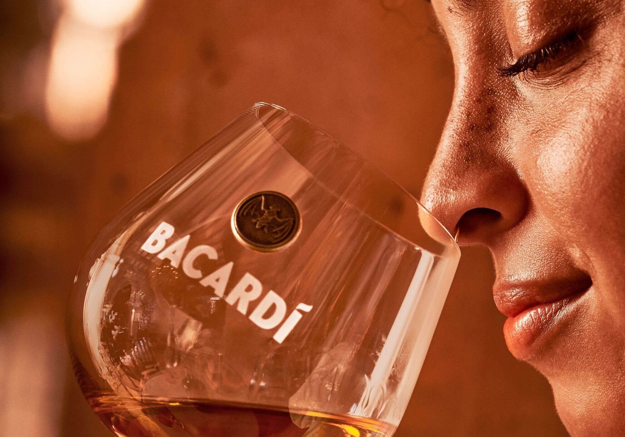 Close-up of a person savoring an amber liquid, bringing a glass with BACARDÍ logo close to their nose.