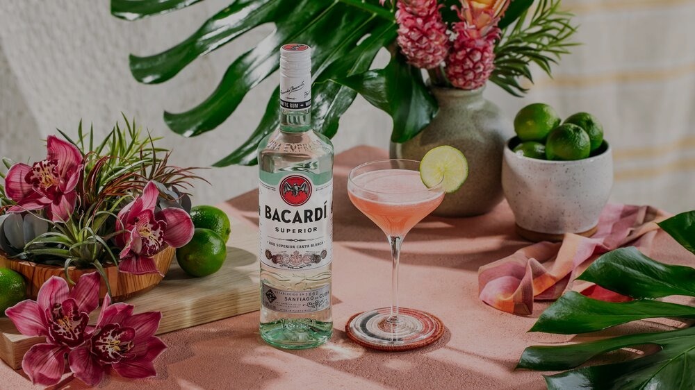 BACARDÍ Superior rum bottle next to pink cocktail, surrounded by limes, orchids, tropical leaves.