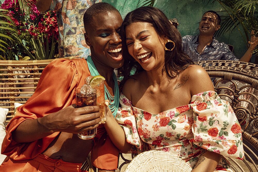 Two people seated together, holding cocktails in an outdoor tropical setting.