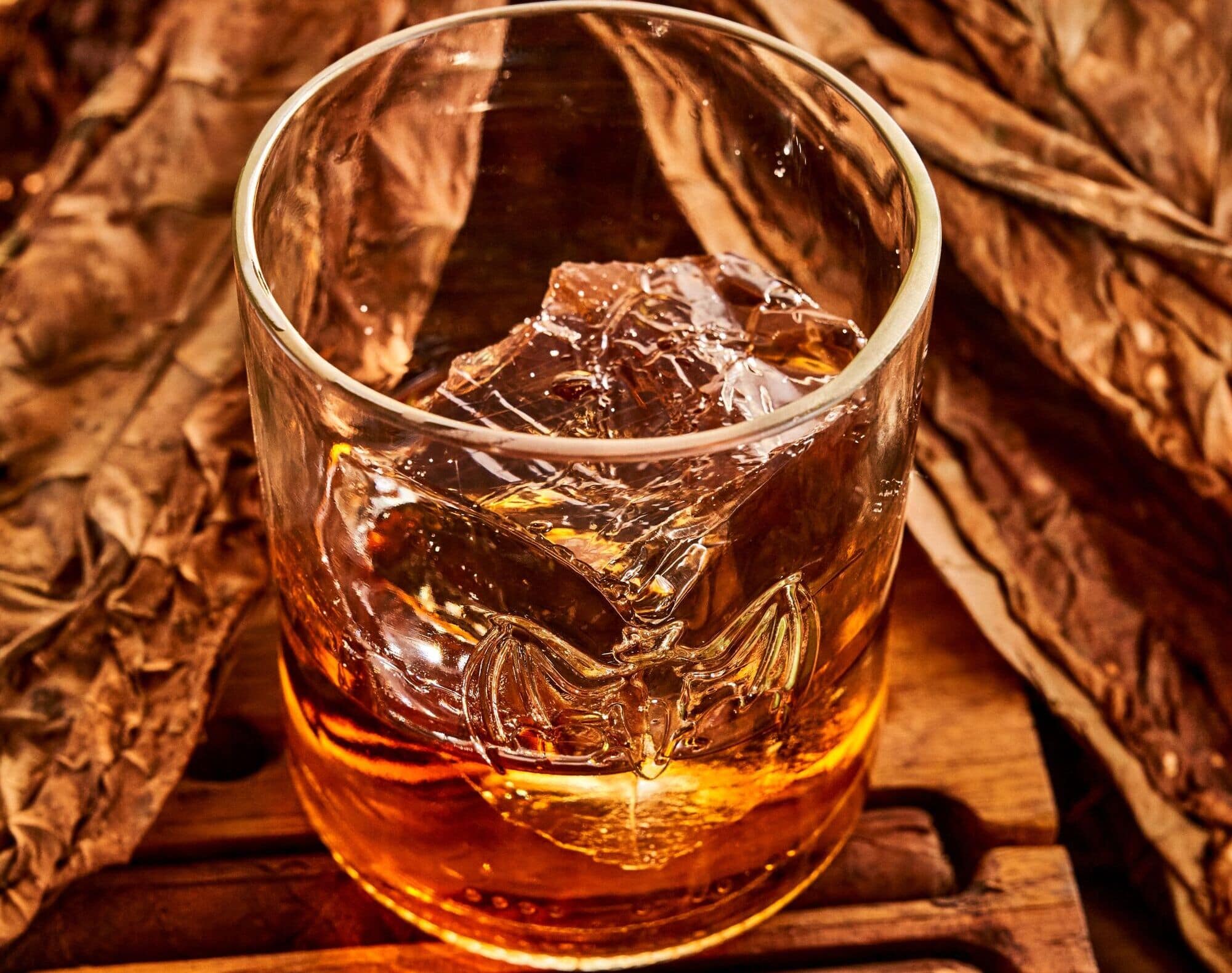 Glass of amber spirit with ice cubes, BACARDÍ bat logo embossed on side, resting near dried tobacco leaves.