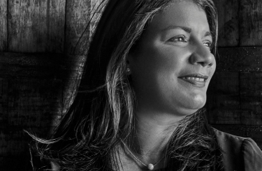 Black and white portrait of a person with long hair, wearing a pearl necklace, looking gently to the right.