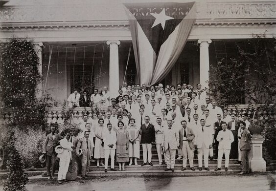 Large group of formally dressed people gathered on building steps and balcony, with Cuban flag draped overhead.