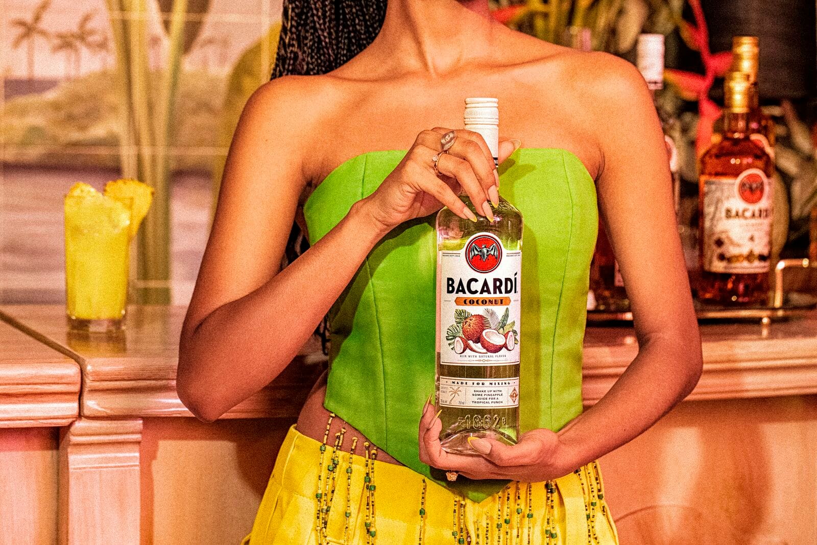 Person holding a BACARDÍ Coconut rum bottle with a cocktail and pineapple garnish nearby.