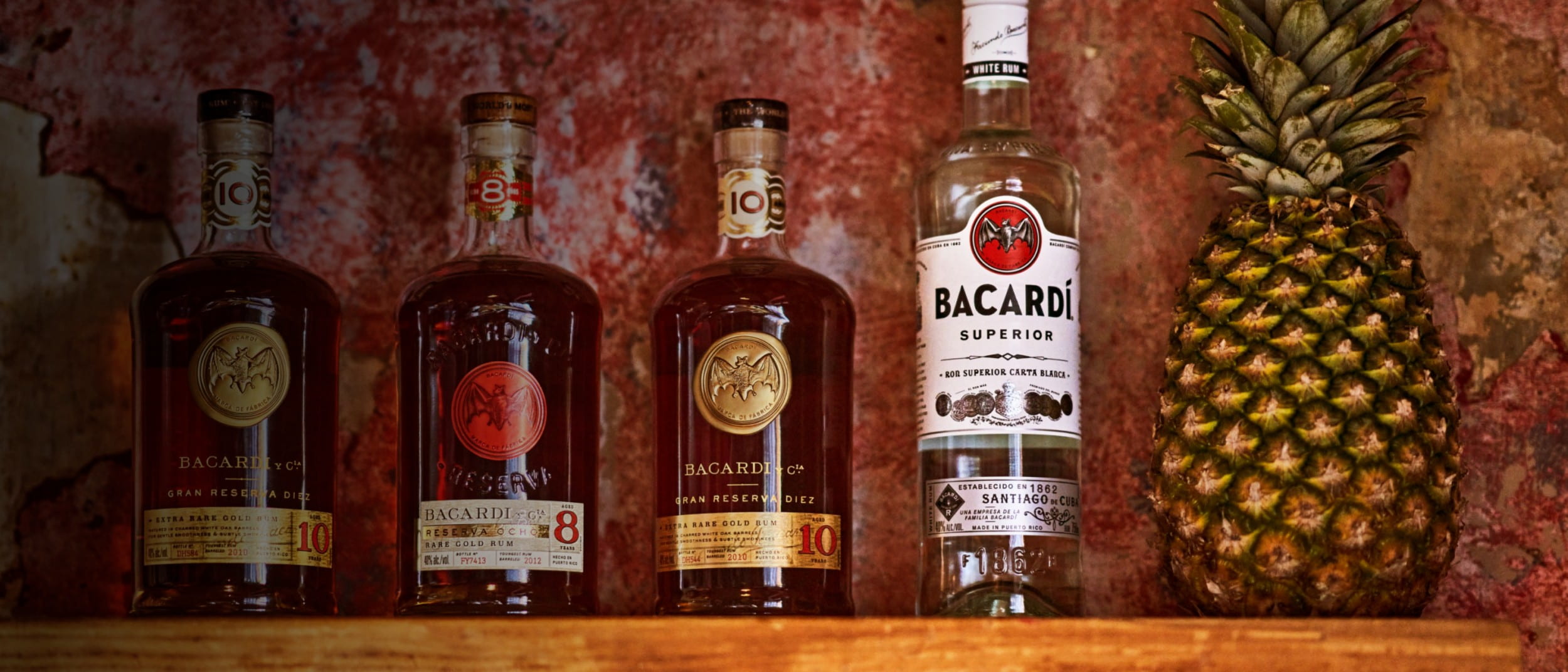 Four BACARDÍ rum bottles in different shades, arranged beside a whole pineapple.