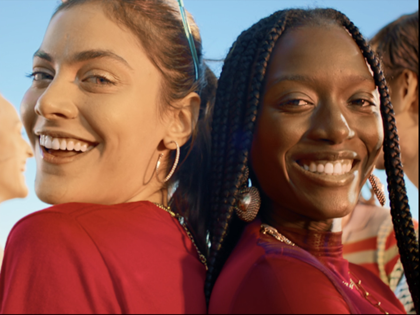 Two people leaning back-to-back, smiling in warm sunlight with other figures nearby.