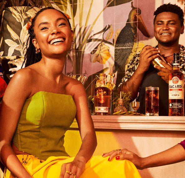 Two people at a vibrant bar with Bacardí rum bottles, cocktail shaker, and tropical mural.